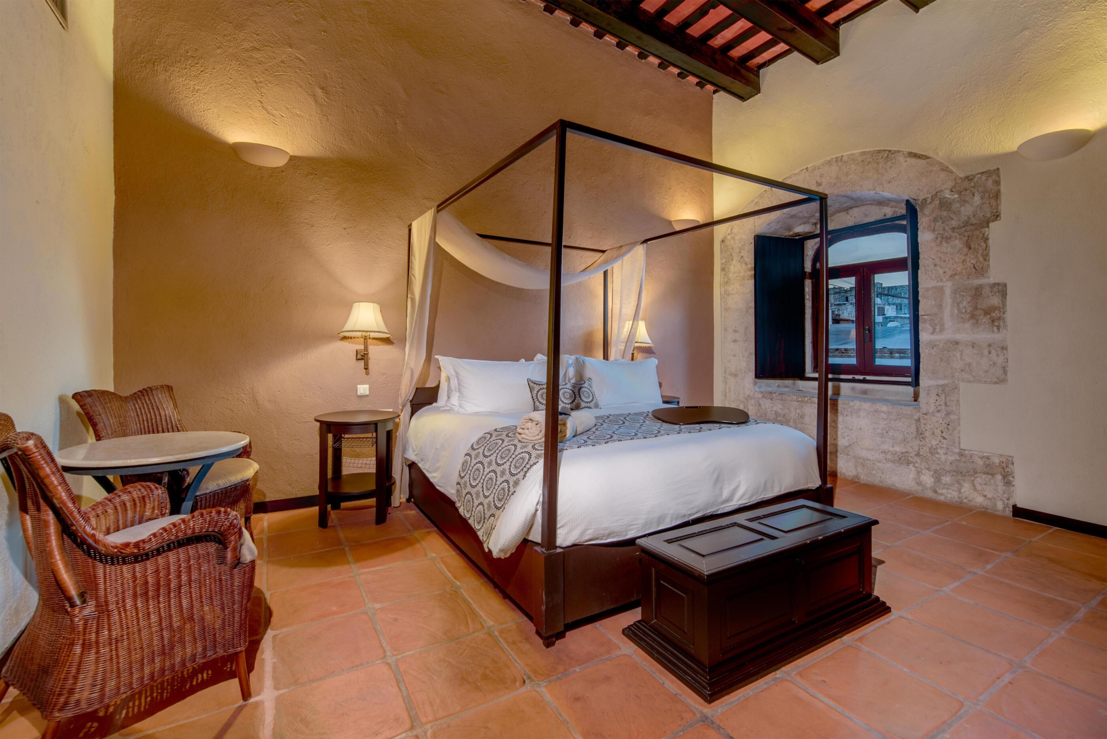 Uma cama com dossel e cortina, uma mesa redonda, cadeiras de vime, uma mesinha de cabeceira e vista para o exterior através de uma janela.
