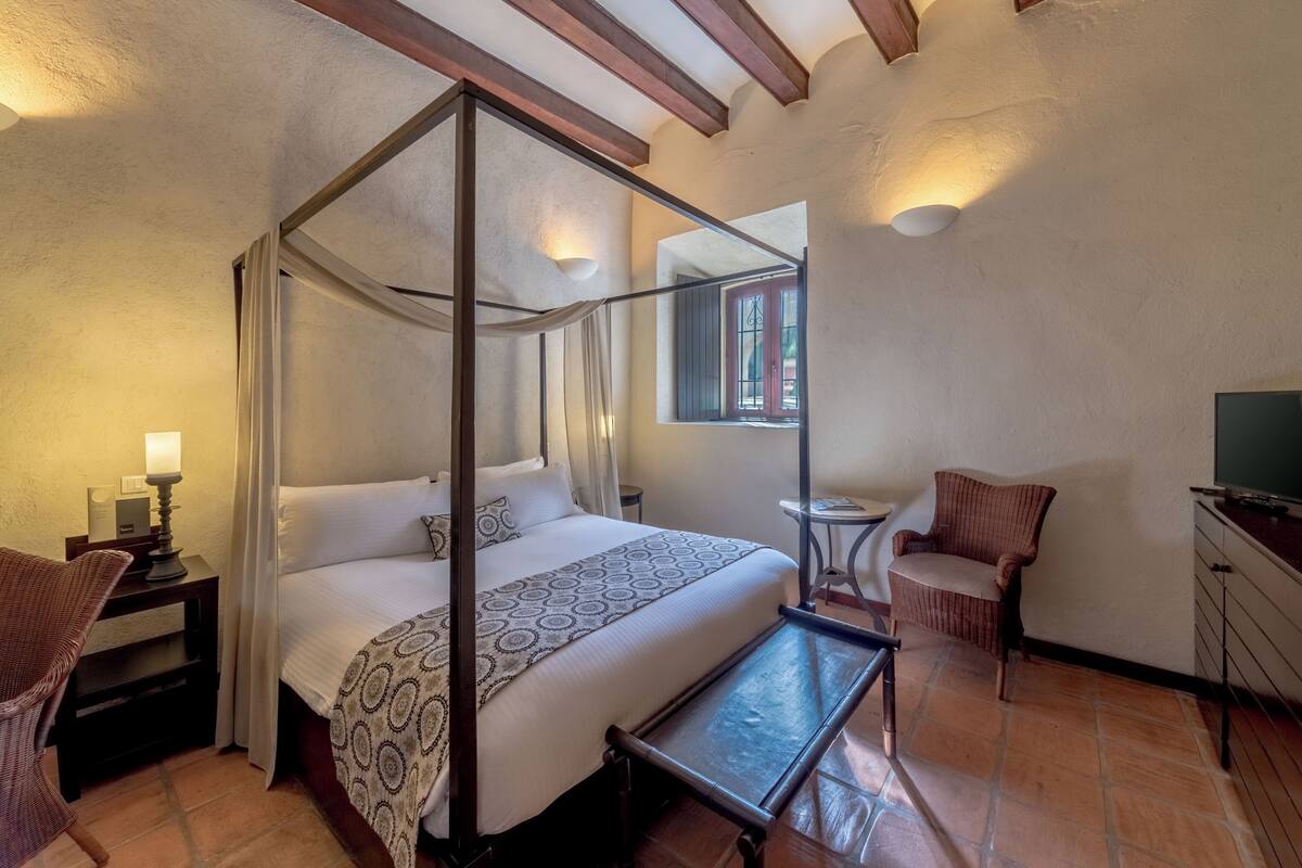 Una cama con dosel, una mesita, una silla y un escritorio con lámpara.