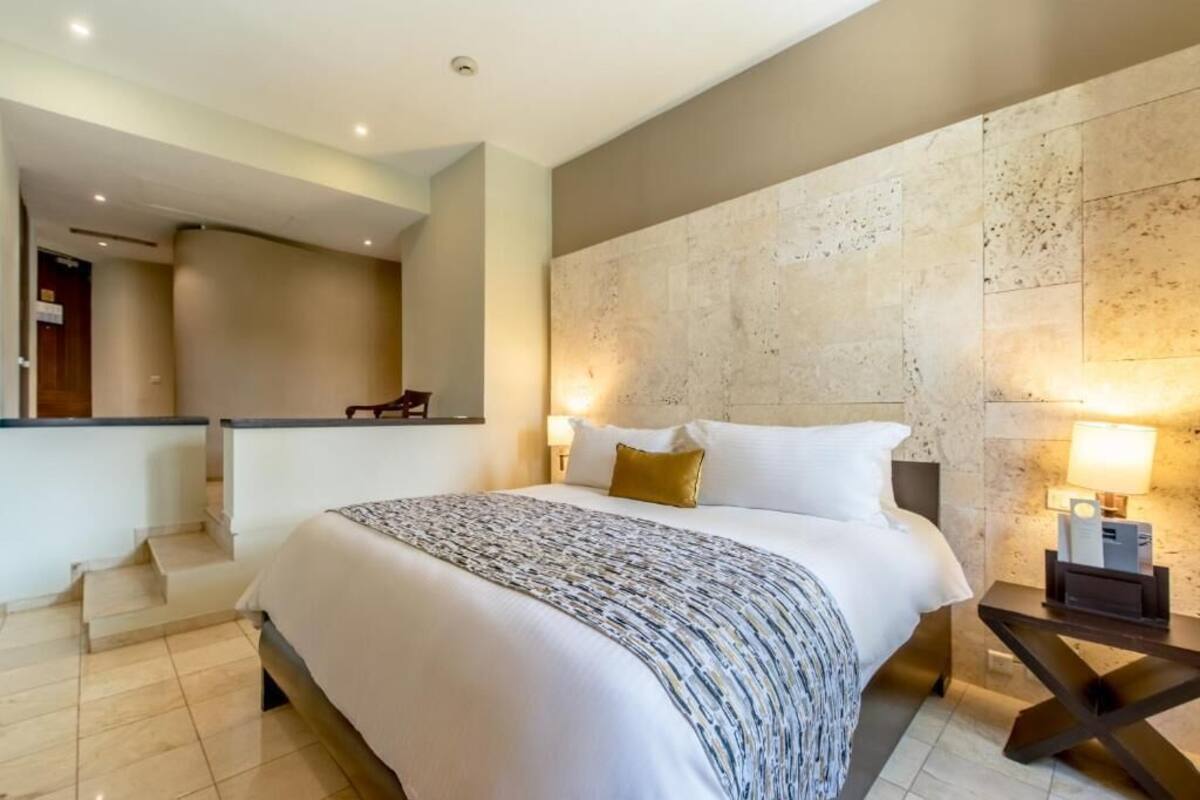 Una habitación de hotel moderna con una cama grande, un cabecero con pared texturizada, una mesita de noche con lámpara y una escalera que lleva a un nivel superior.