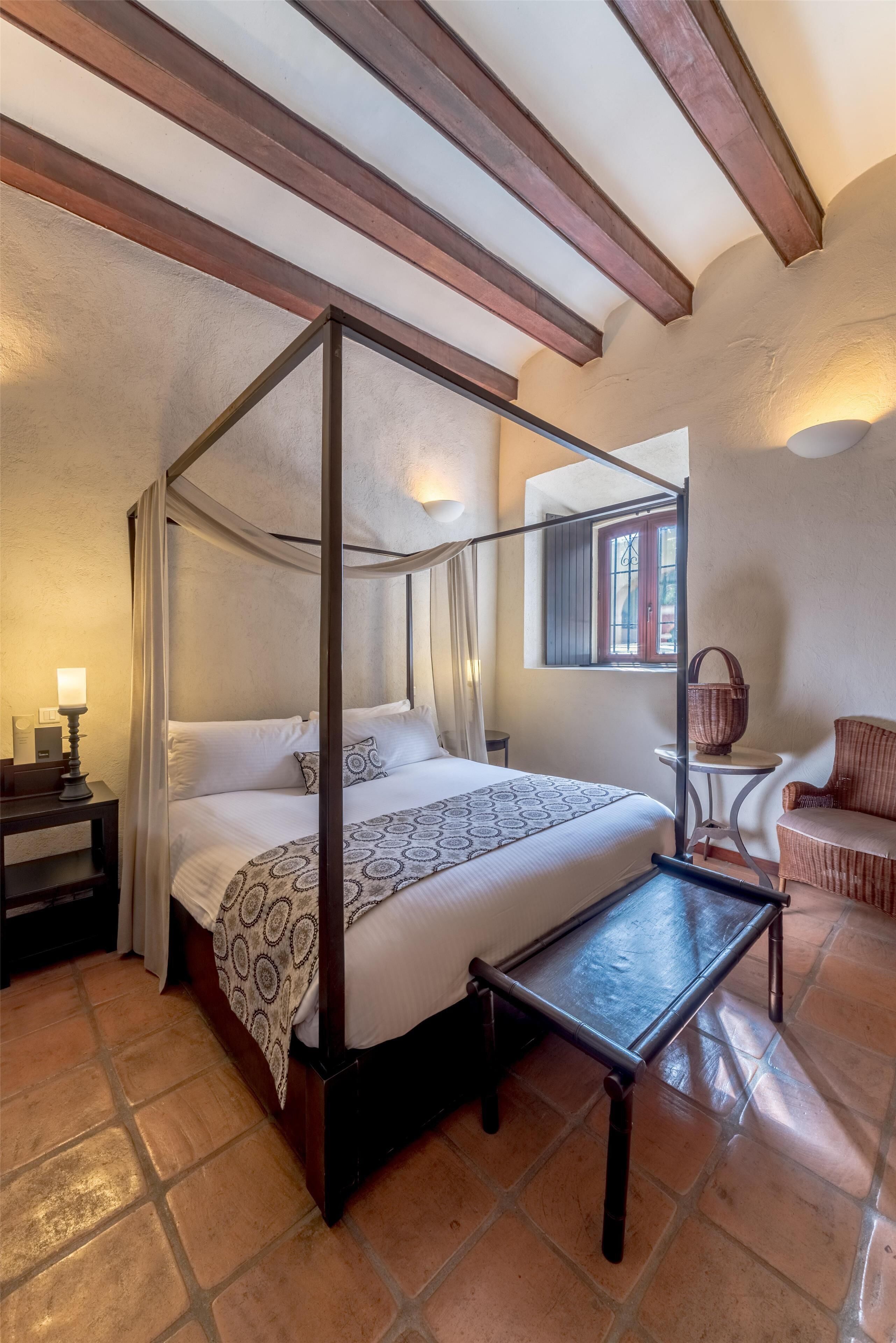 Una cama con dosel, una mesita, un sillón de mimbre y una mesita de noche con lámpara.