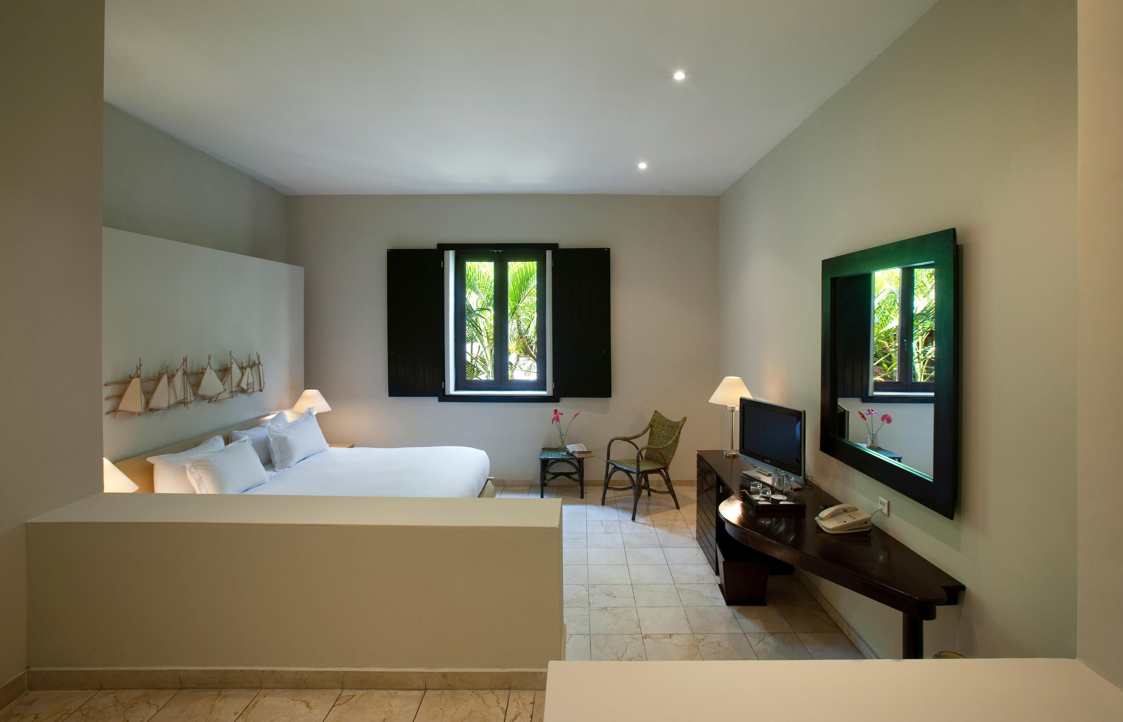 A hotel room with a bed, a desk with a phone, a chair, and two windows with shutters.
