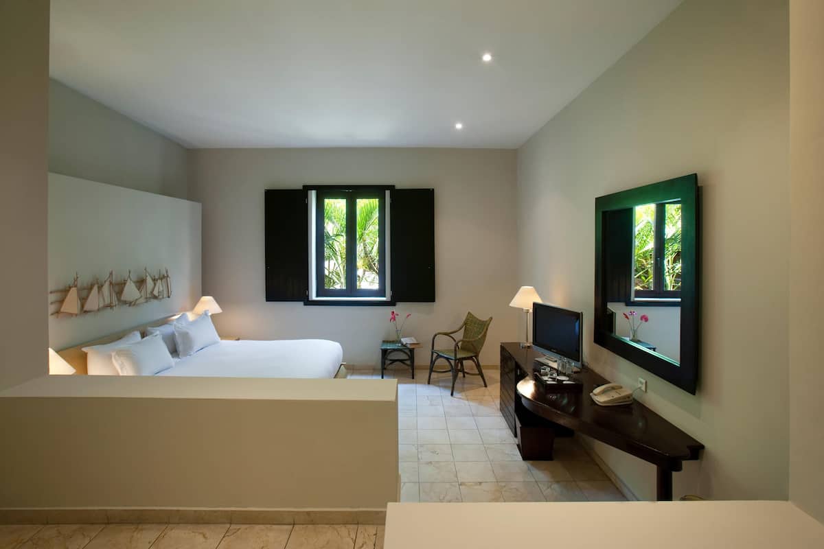 A hotel room with a bed, a desk with a phone, a chair, and two windows with shutters.