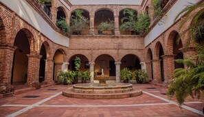 Une cour intérieure avec une fontaine centrale, entourée d’arcades en briques et de plantes en pot.