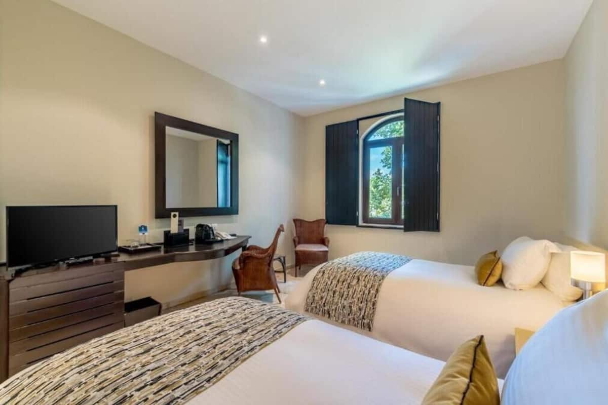 A hotel room with two beds, a desk with a TV, a chair, a window with curtains, and a mirror on the wall.