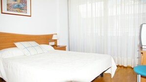 A neatly made bed with a white bedspread and a blue pillow, a wooden nightstand with a lamp, and a large window with sheer curtains. - Apartamento Ancora (Tossa de Mar)