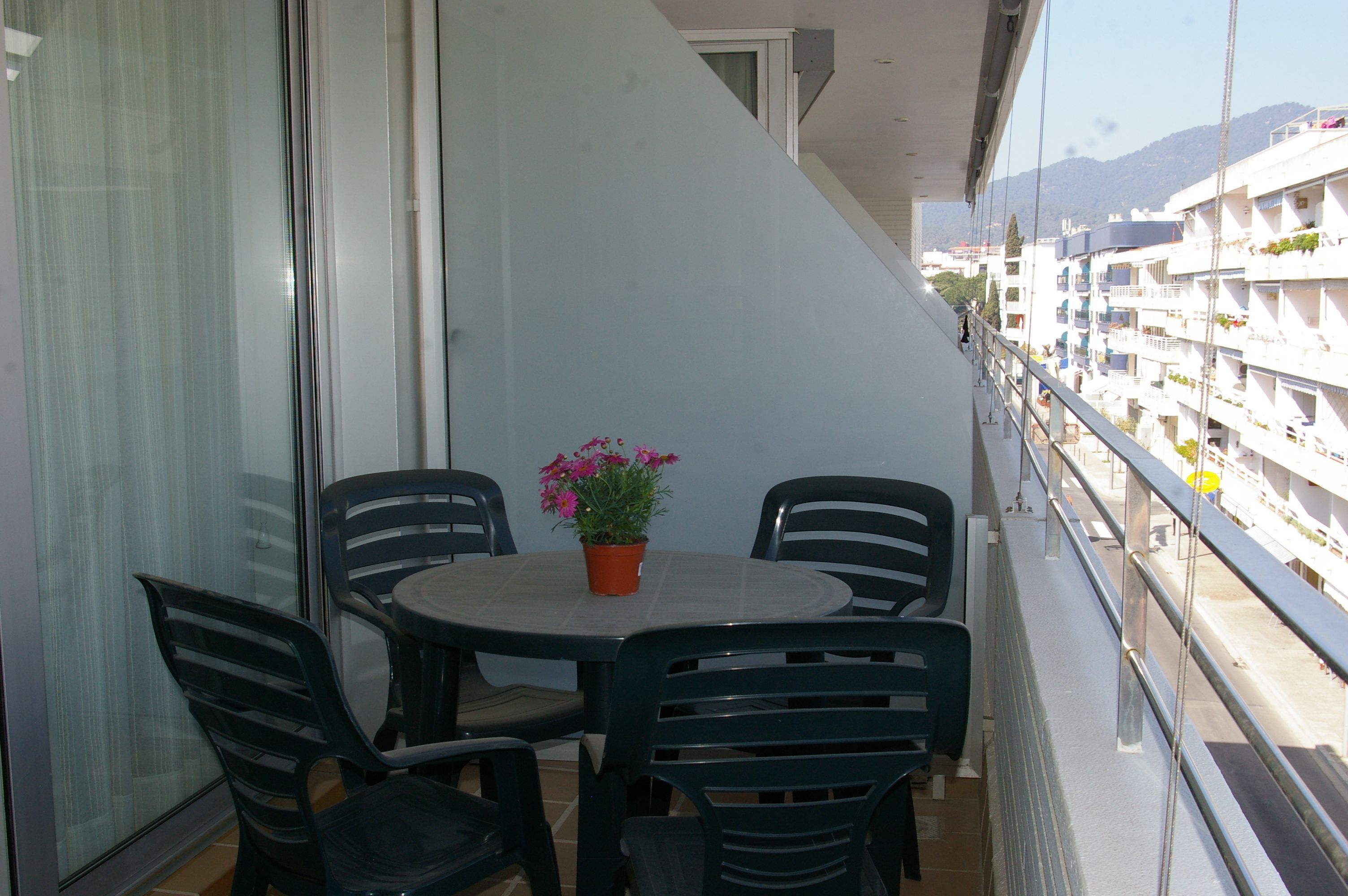 Un balcon avec une table et des chaises, un pot de fleurs et une vue sur un quartier résidentiel, avec des bâtiments et des montagnes en arrière-plan.