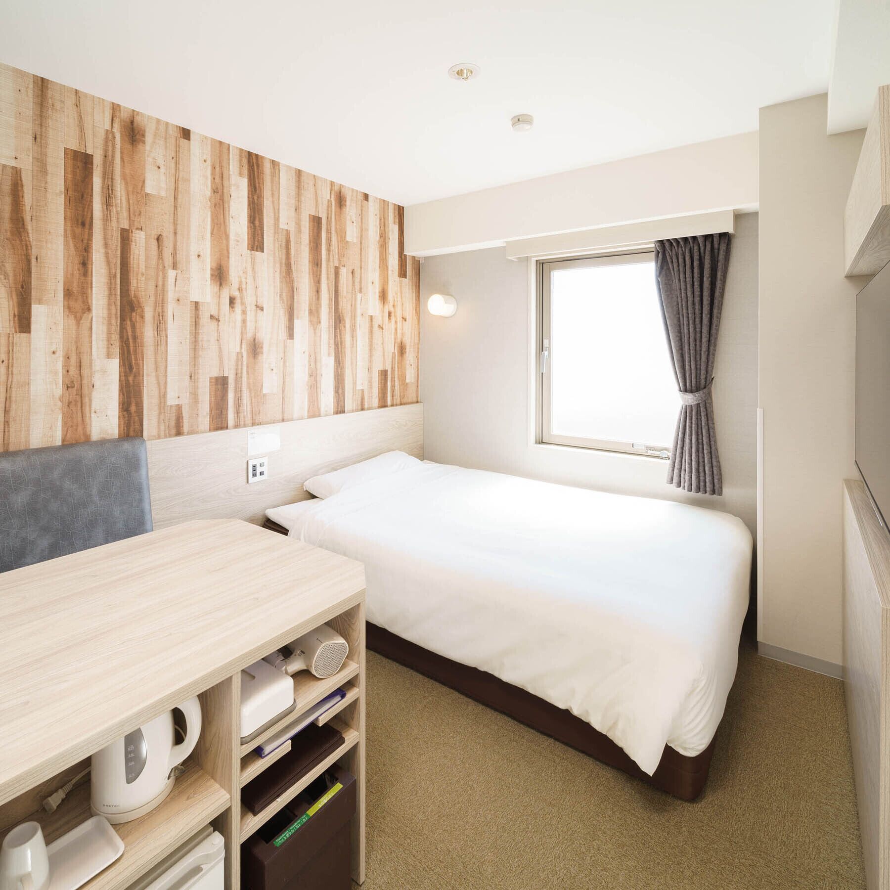 A compact hotel room with a bed, a desk, a chair, a window with curtains, and a wooden wall panel.