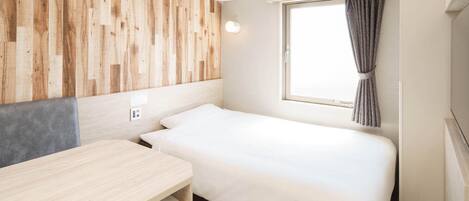 A compact hotel room with a bed, a desk, a chair, a window with curtains, and a wooden wall panel.