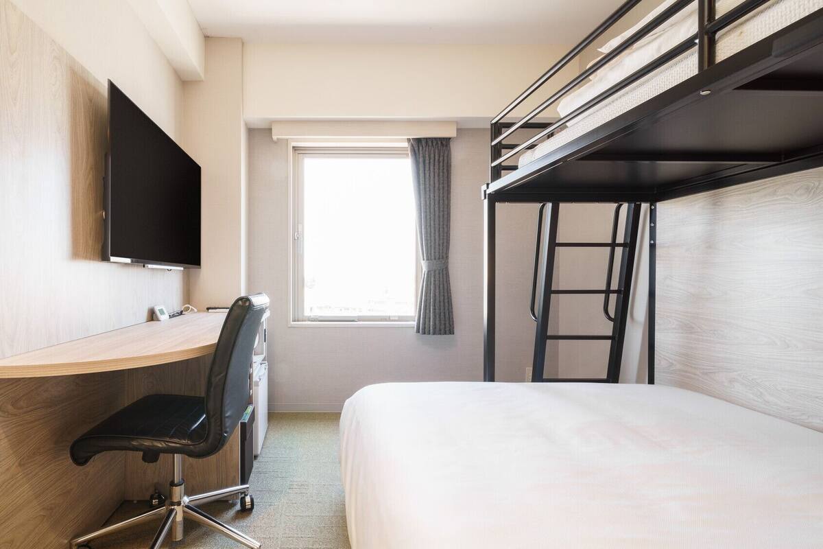 Une chambre d’hôtel compacte comprenant un lit superposé, un bureau avec une chaise, une télévision et une fenêtre avec des rideaux.