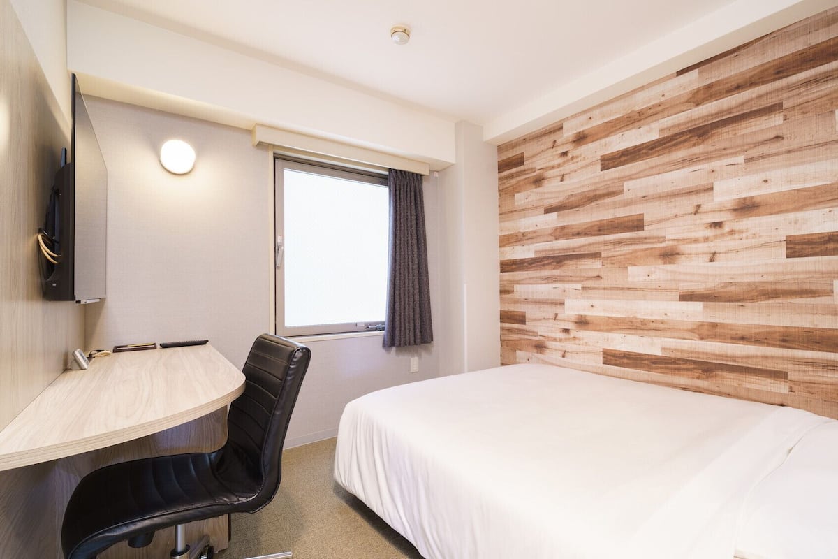Hotellihuone, jossa on sänky, työpöytä, tuoli, ikkuna verhoilla ja puinen seinäpaneeli.