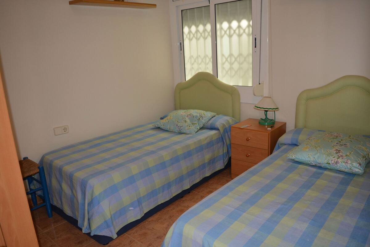 Una habitació amb dos llits individuals, cadascun amb almoina estampada amb motius florals i un llençol de quadres. Hi ha una tauleta de nit amb una làmpada entre els llits.