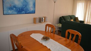 Un comedor con una mesa de madera, sillas, un camino de mesa blanco, un cuadro abstracto azul, un sofá verde, una lámpara y un altavoz.