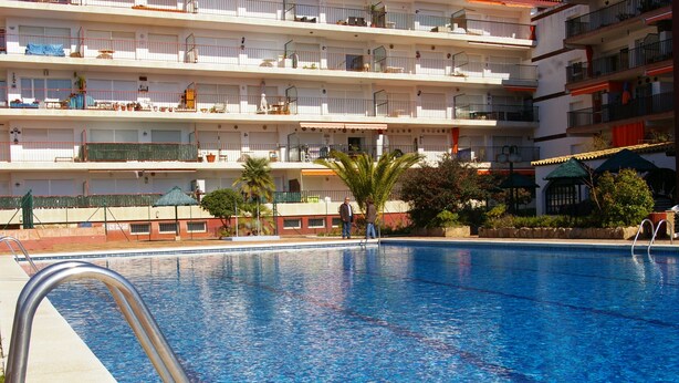 Piscina con una escalera metálica, rodeada por un edificio con balcones, palmeras y mobiliario exterior.