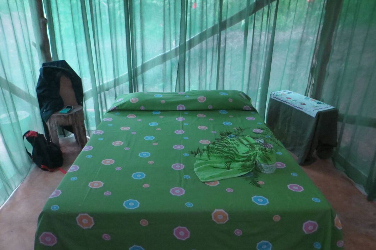 A room with a bed covered in green bedding with a polka dot pattern, a wooden stool, and a black bag on the floor.