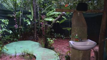 A rustic bathroom with a sink and a mirror, surrounded by dense foliage and a green mat.