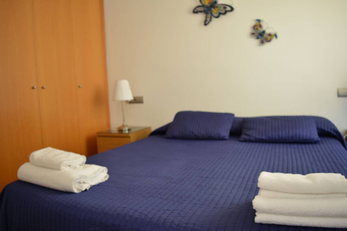 A neatly made bed with blue bedding and pillows, a wooden wardrobe, and a nightstand with a lamp.