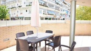 An outdoor patio with a table and chairs, a covered umbrella, and a view of a hotel building in the background. - Apartamento Voramar (Tossa de Mar)