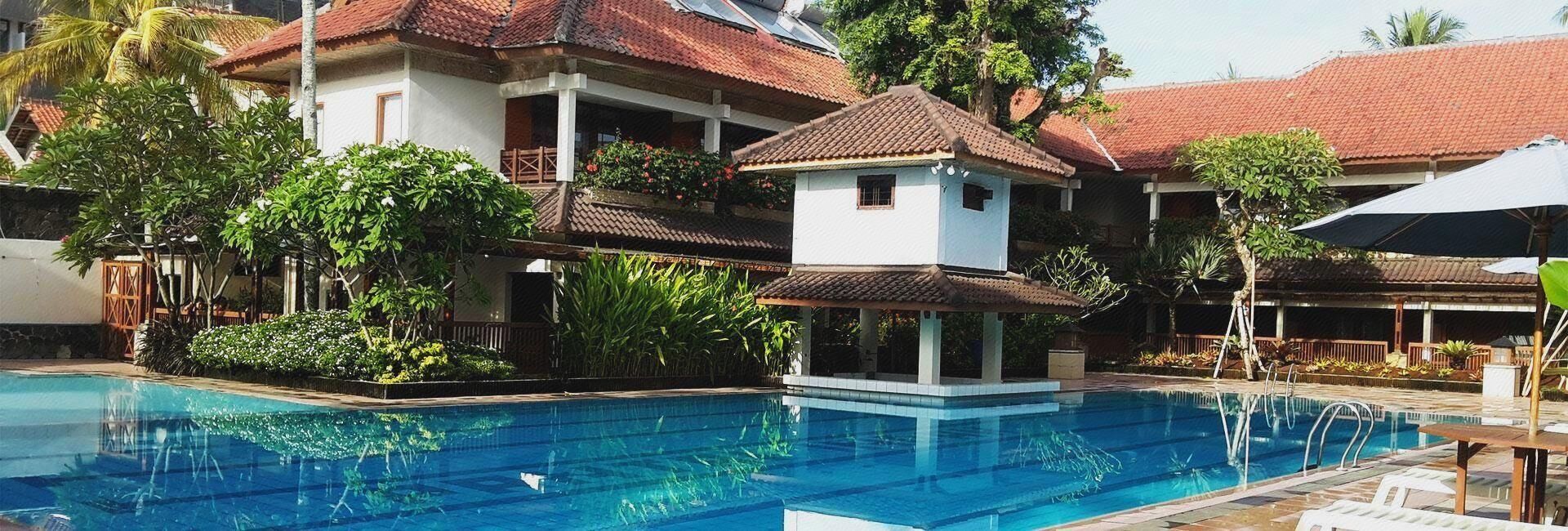 En swimmingpool med en liten paviljong, omgiven av frodig grönska och traditionell arkitektur.
