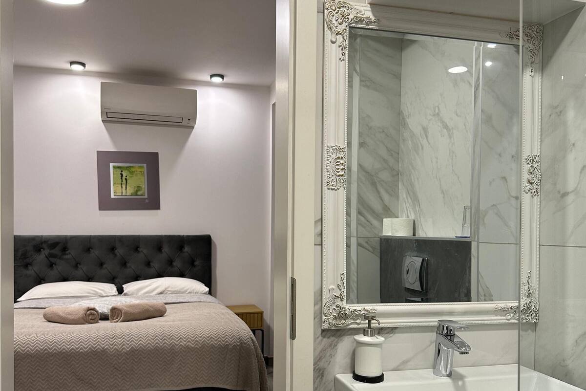 A bedroom with a bed, a bedside table, a wall-mounted air conditioner, and a framed picture.