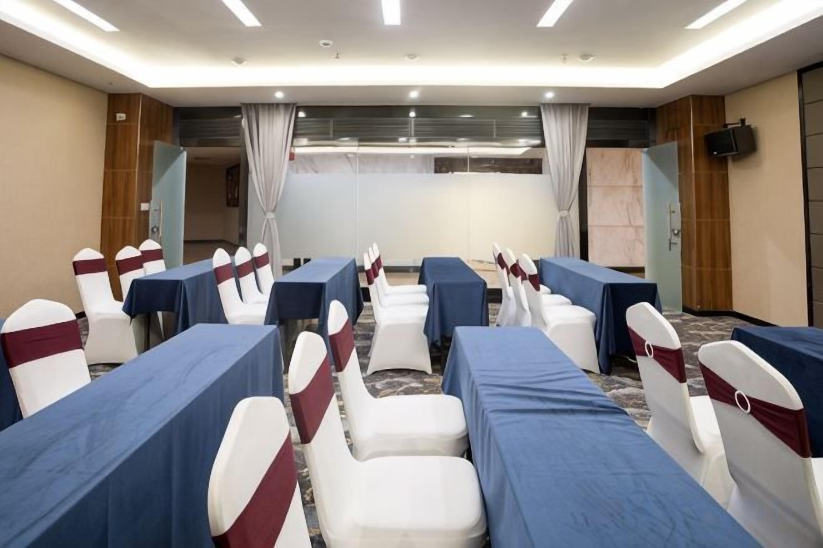 A conference room with rows of tables and chairs, a large screen at the front, and a wooden panel wall.