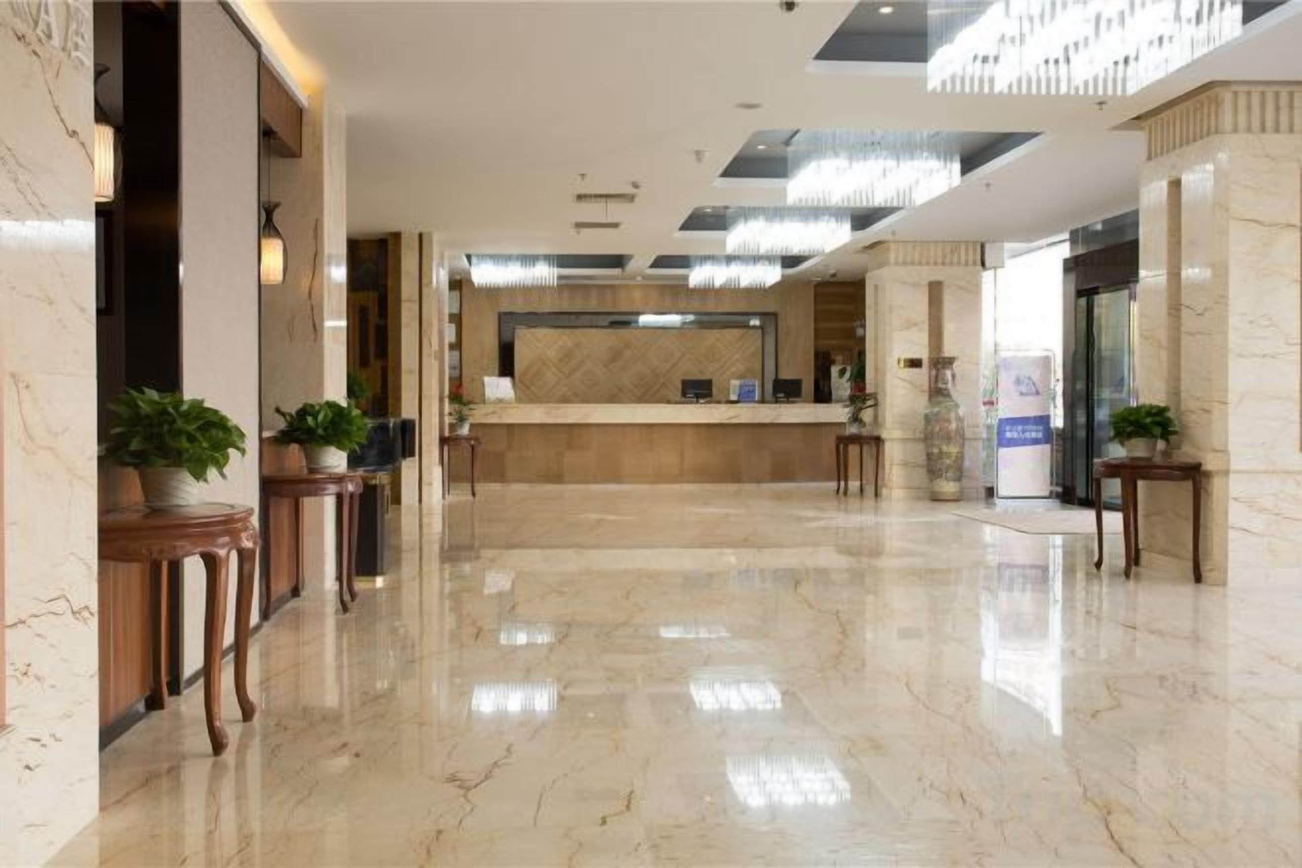 A hotel lobby with a reception desk, marble flooring, and potted plants.