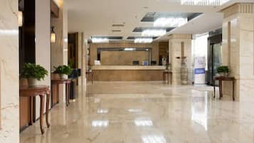 A hotel lobby with a reception desk, marble flooring, and potted plants.