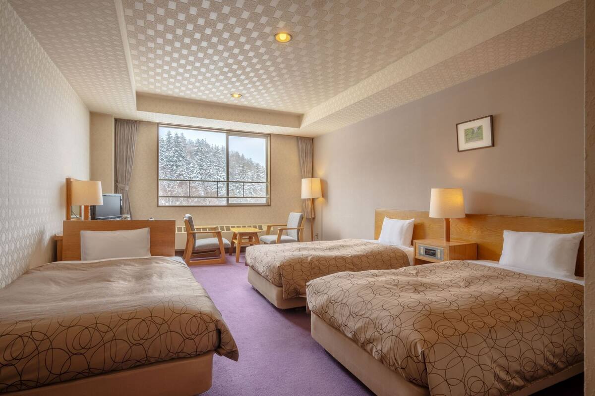 Ein Hotelzimmer mit zwei Betten, einem Schreibtisch, zwei Stühlen, einem Fenster mit Blick auf schneebedeckte Bäume und einer Decke mit einem gemusterten Design.