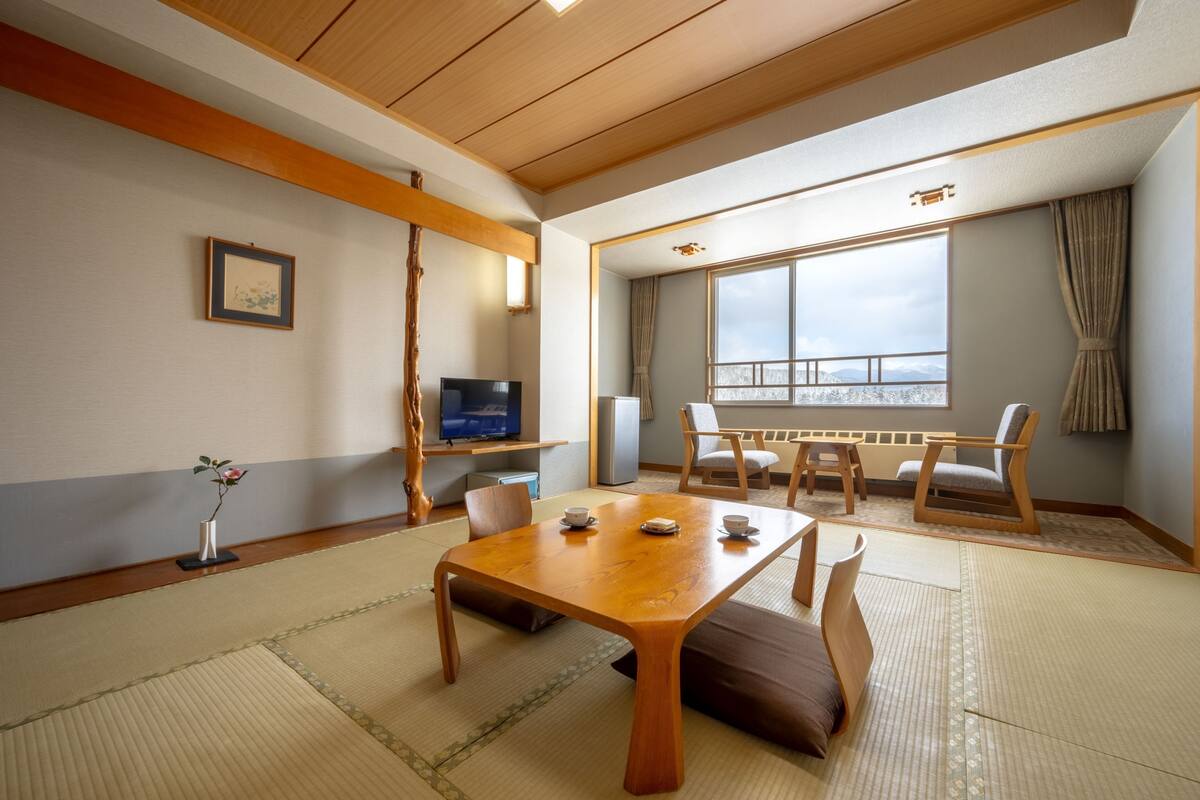 Ein traditioneller japanischer Raum mit Tatami-Boden, einem Holztisch mit Kissen, einem Fernseher und einem großen Fenster mit Aussicht.