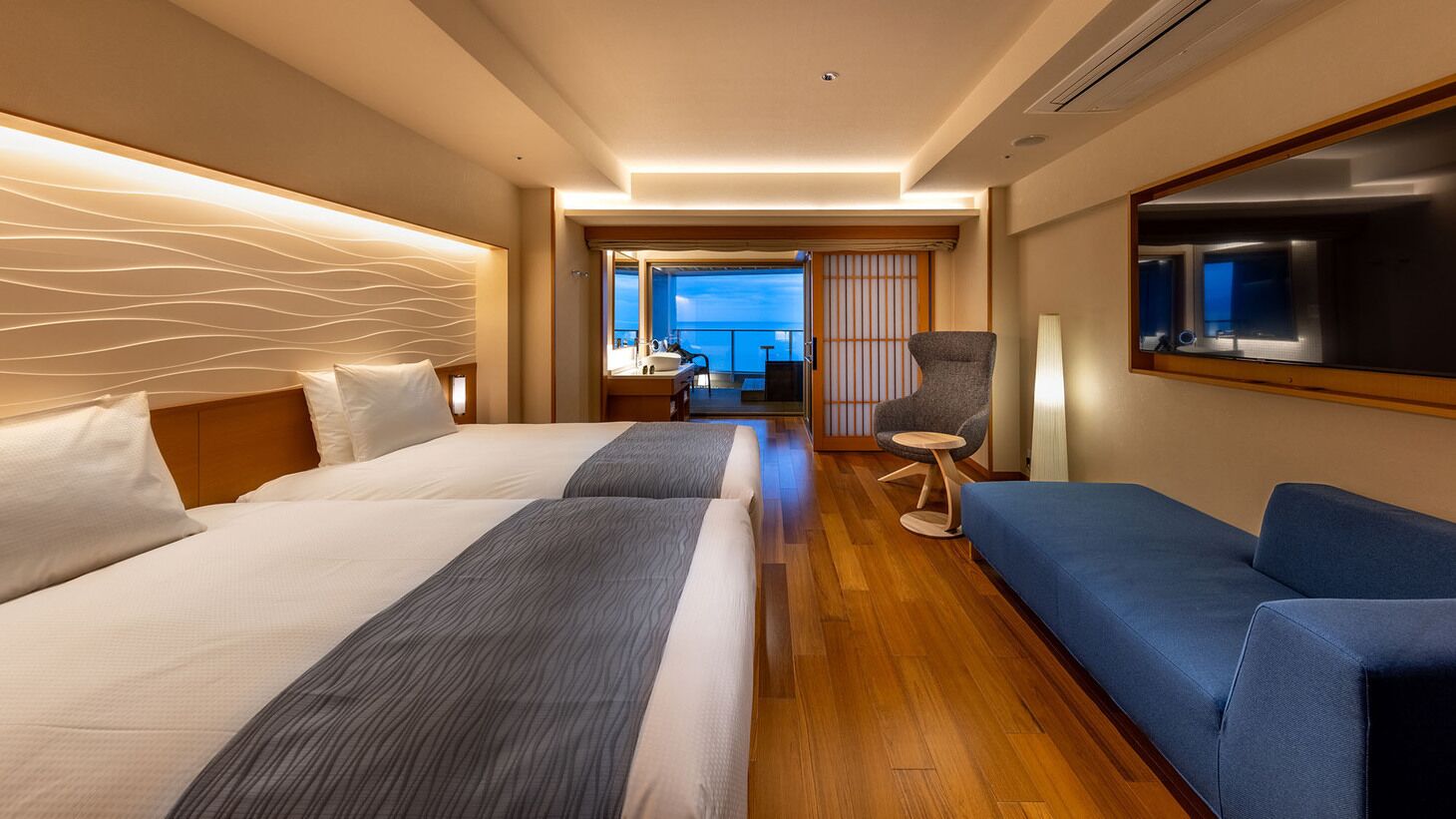 A hotel room with two beds, a sofa, a chair, a TV, and a view of the sea.