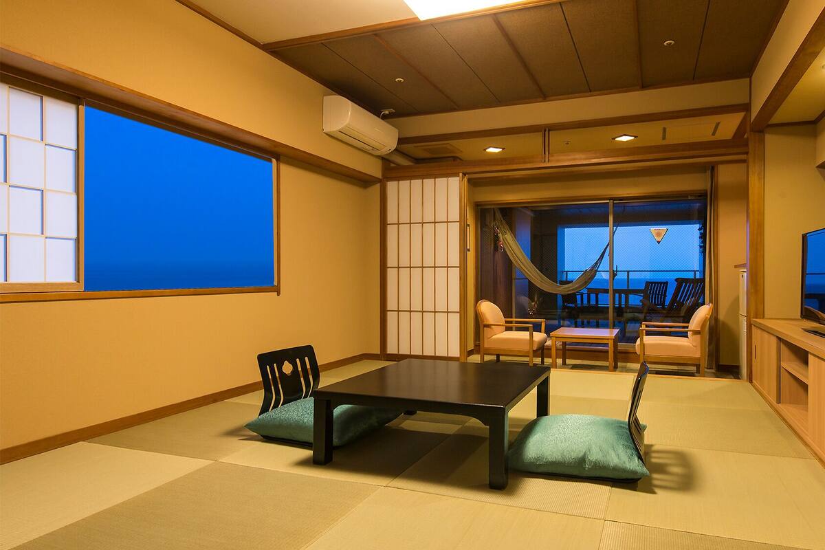 Ein traditioneller japanischer Raum mit Tatami-Boden, einem niedrigen Tisch, Stühlen, einem Fernseher und Blick durch eine Schiebetür.
