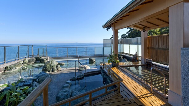 Una terraza de madera con barandilla de vidrio, con vistas a una piscina con rocas y otra terraza de madera.