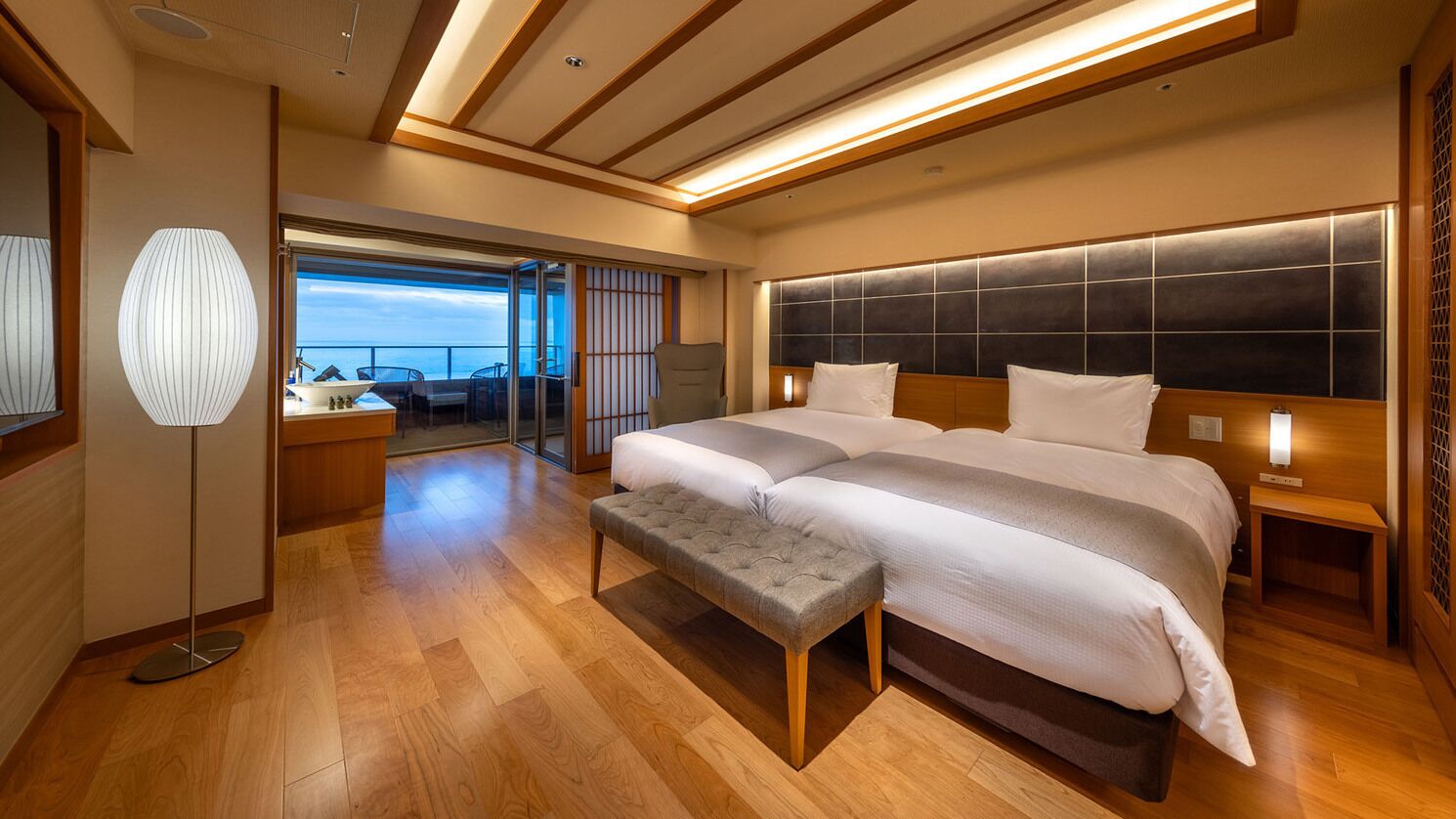 A modern hotel room with a large bed, a bench, a bedside table, and a view of the sea.