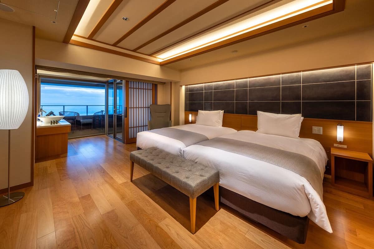Una habitación de hotel moderna con una cama grande, un banco, una mesita de noche y vistas al mar.