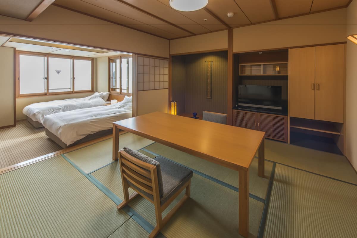 Ein traditioneller japanischer Raum mit Tatami-Böden, einem niedrigen Holztisch, einem Bett mit weißer Bettwäsche, einem Fernseher und gleitenden Türen.