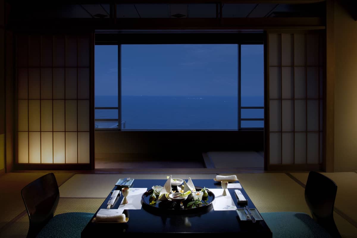 Ein traditioneller japanischer Raum mit Tatami-Böden, einem niedrigen Tisch mit einer Mahlzeit und Blick auf das Meer durch Shoji-Schirme.