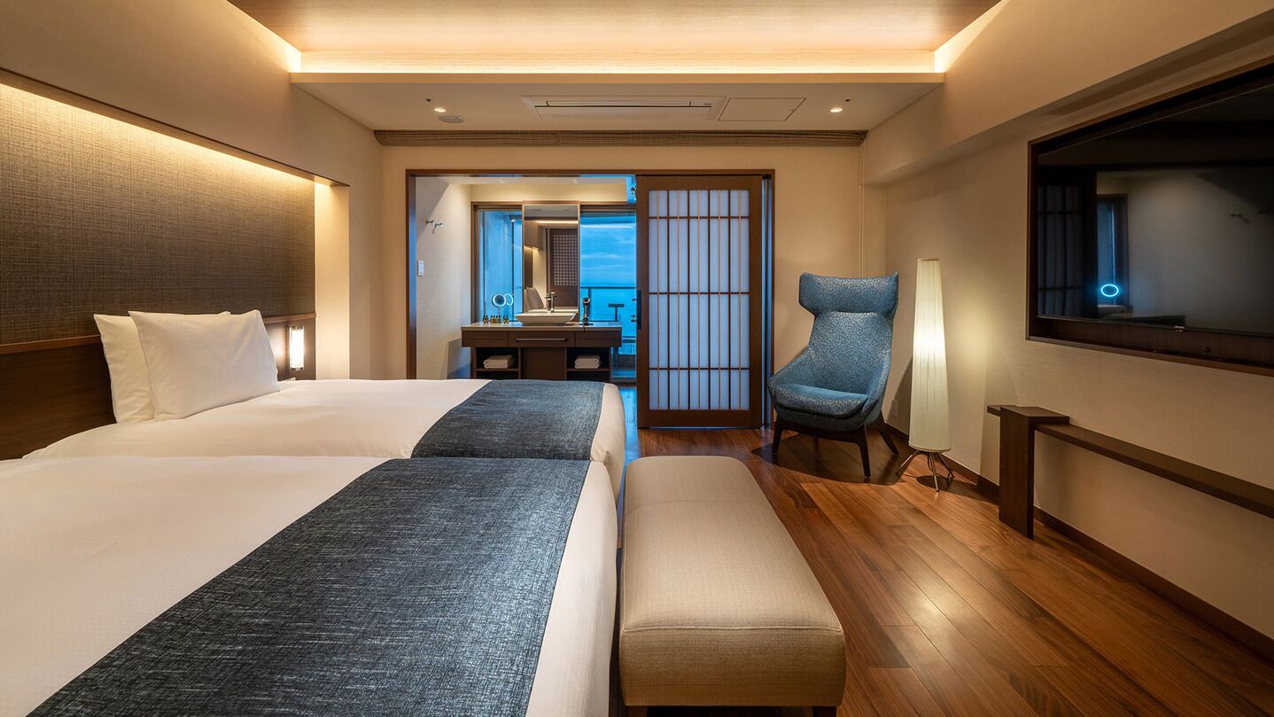 A hotel room with two beds, a desk, a chair, a TV, and a view of the sea.
