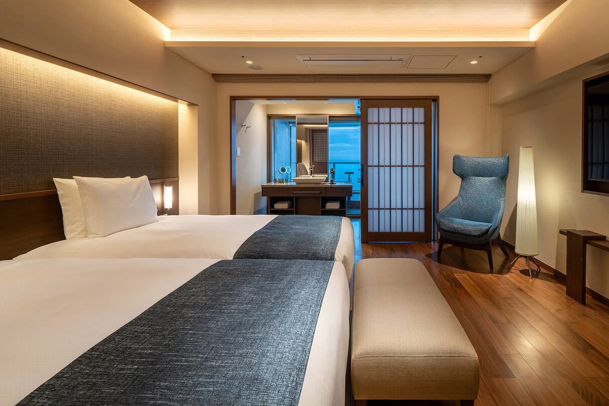 Ein Hotelzimmer mit zwei Betten, einem Schreibtisch, einem Sessel, einem Fernseher und Meerblick.