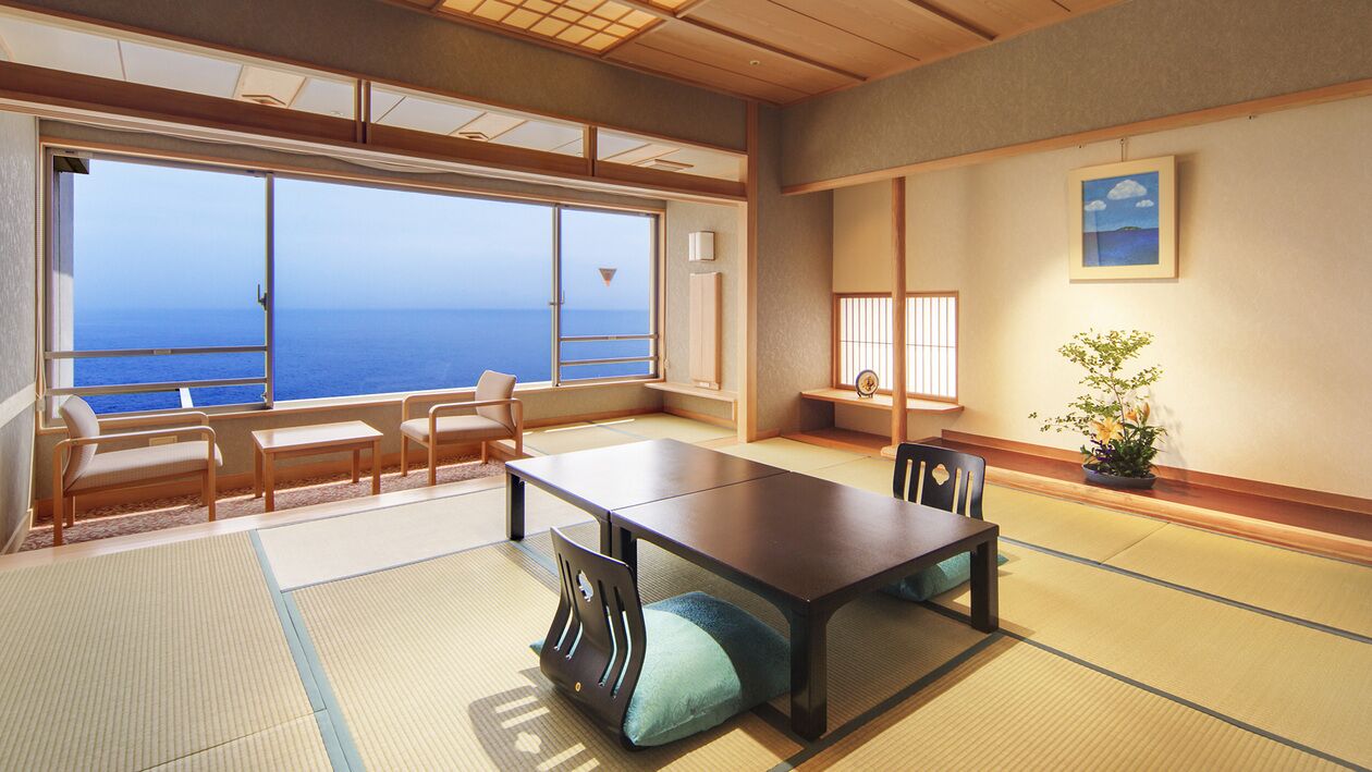 A room with tatami flooring, a low table, chairs, a large window with a view of the sea, and a small plant.
