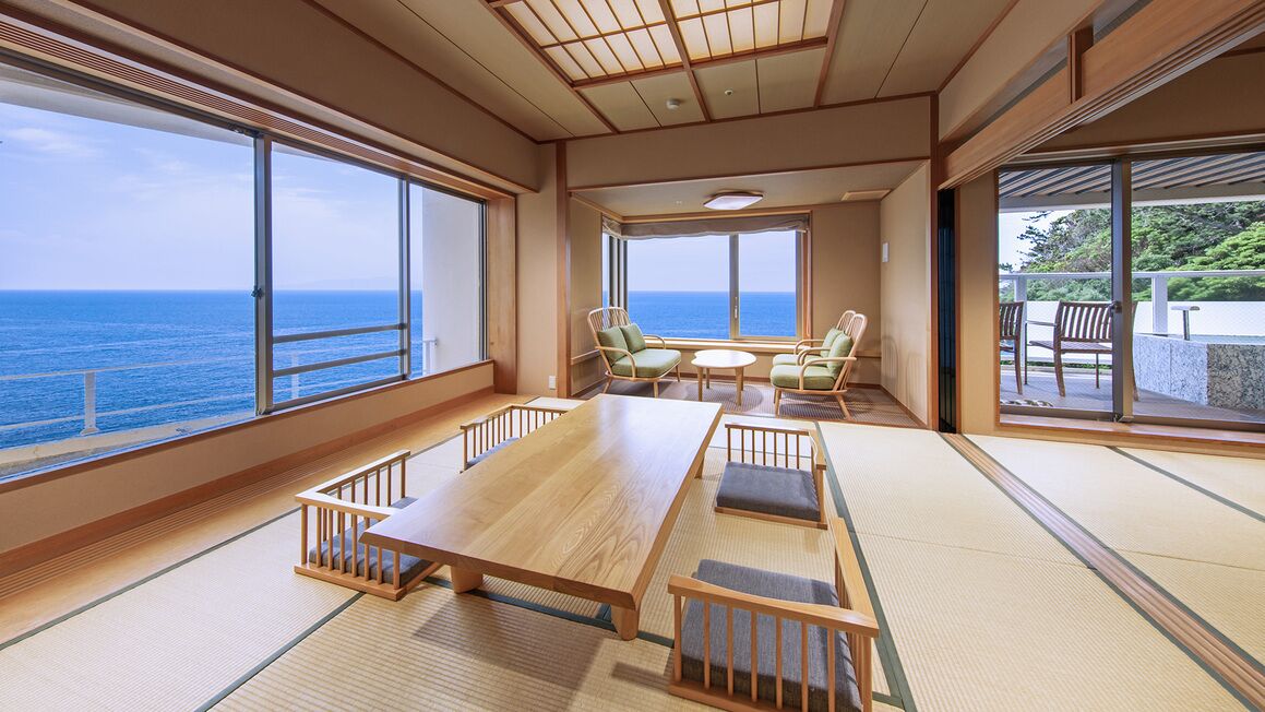 A room with tatami flooring, a wooden table, chairs, and large windows offering a view of the sea.