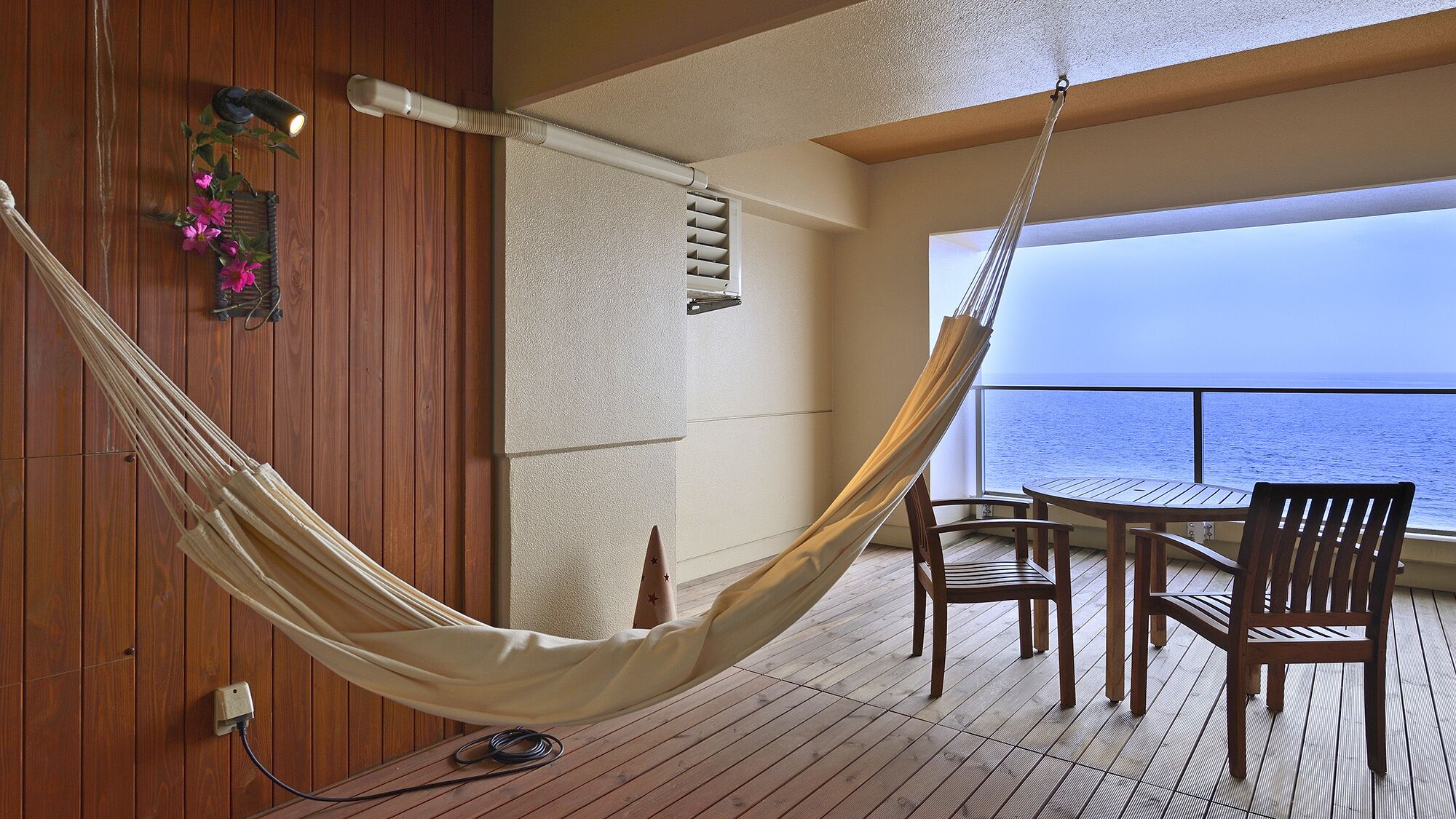 Una hamaca en una terraza de madera con una mesa, sillas, un ventilador de techo y vista al mar.