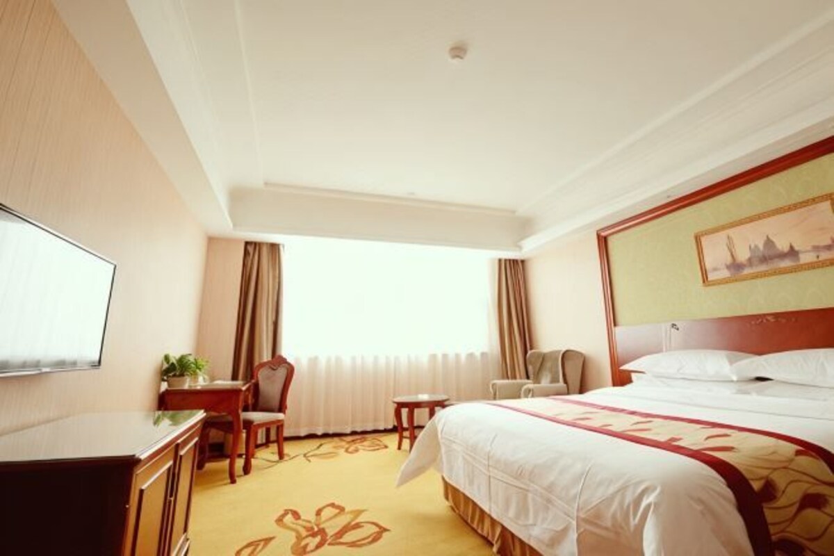 A hotel room with a bed, a desk, a chair, a TV, and a window with curtains.