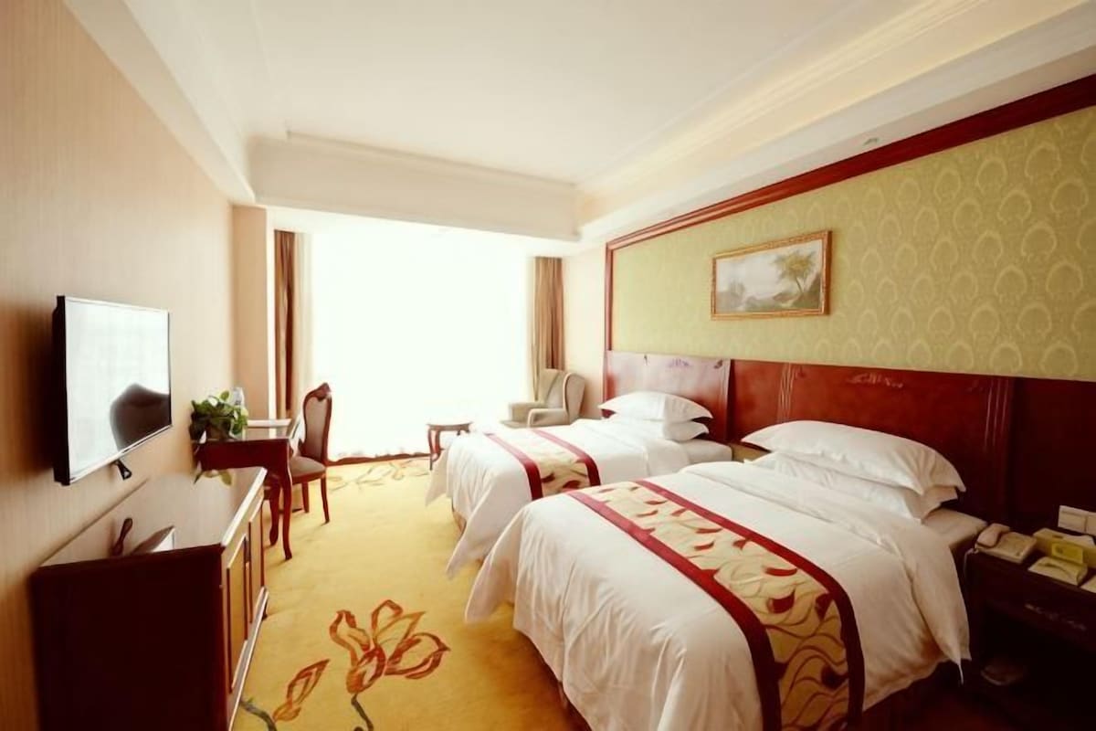 A hotel room with two beds, a desk, a chair, a television, and a window.