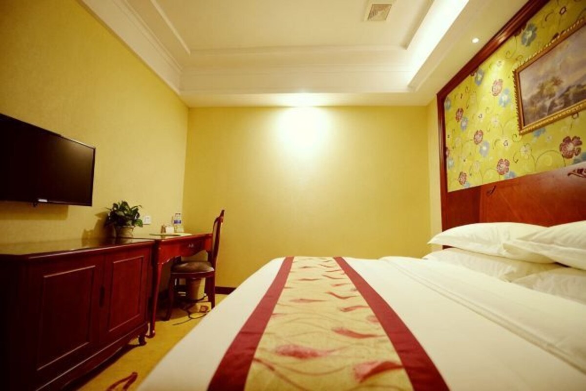 A hotel room with a bed, a desk, a chair, a television, and a painting on the wall.