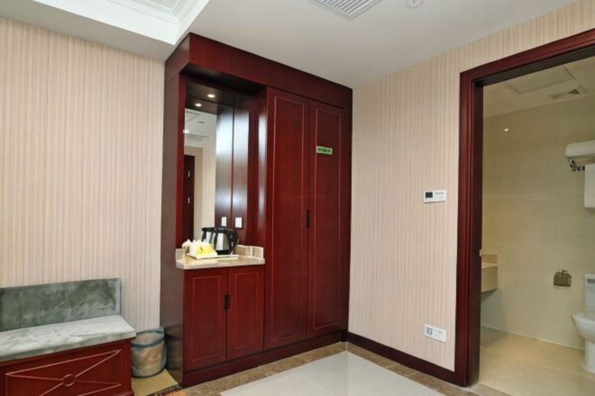 A hotel room with a wooden wardrobe, a bench, a small table with a kettle, and a mirror.