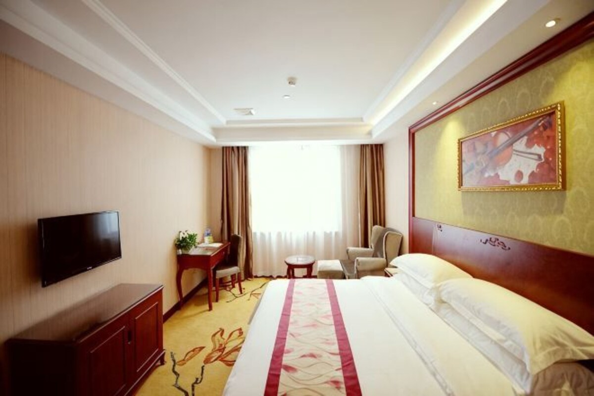 A hotel room with a bed, a television, a desk, a chair, and a painting on the wall.