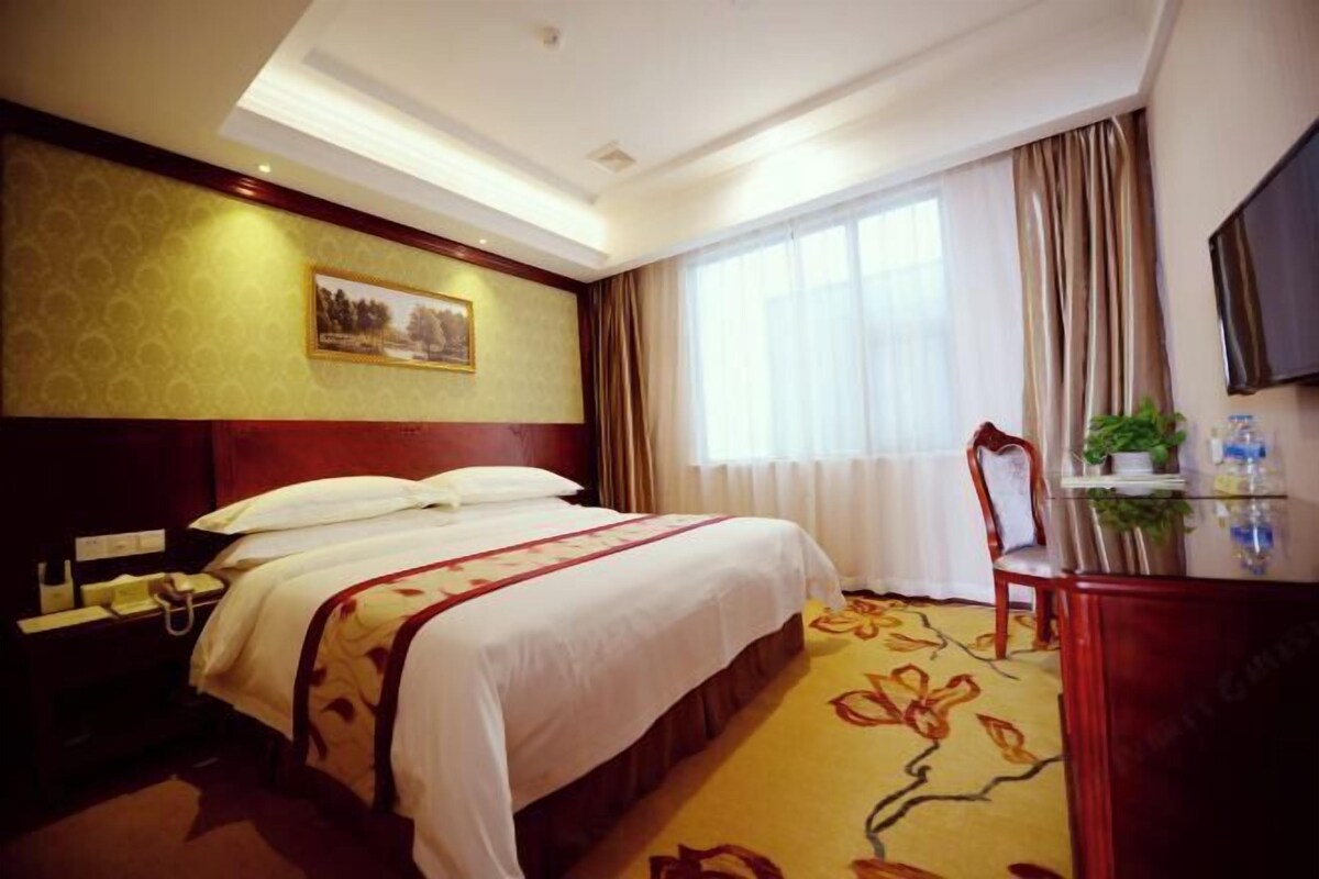 A hotel room with a large bed, a desk with a chair, a television, and a window with curtains.