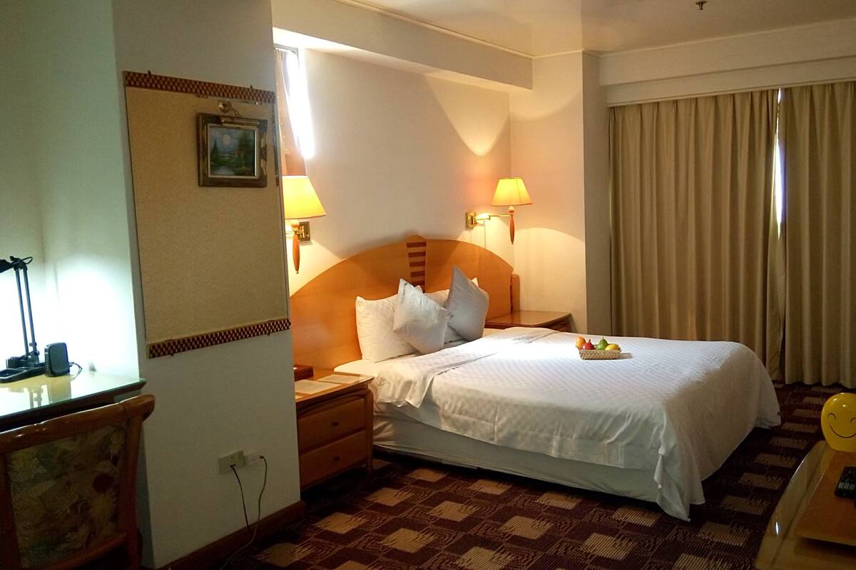 A hotel room with a bed, two bedside lamps, a nightstand, a desk with a lamp, a television, and a window with curtains.