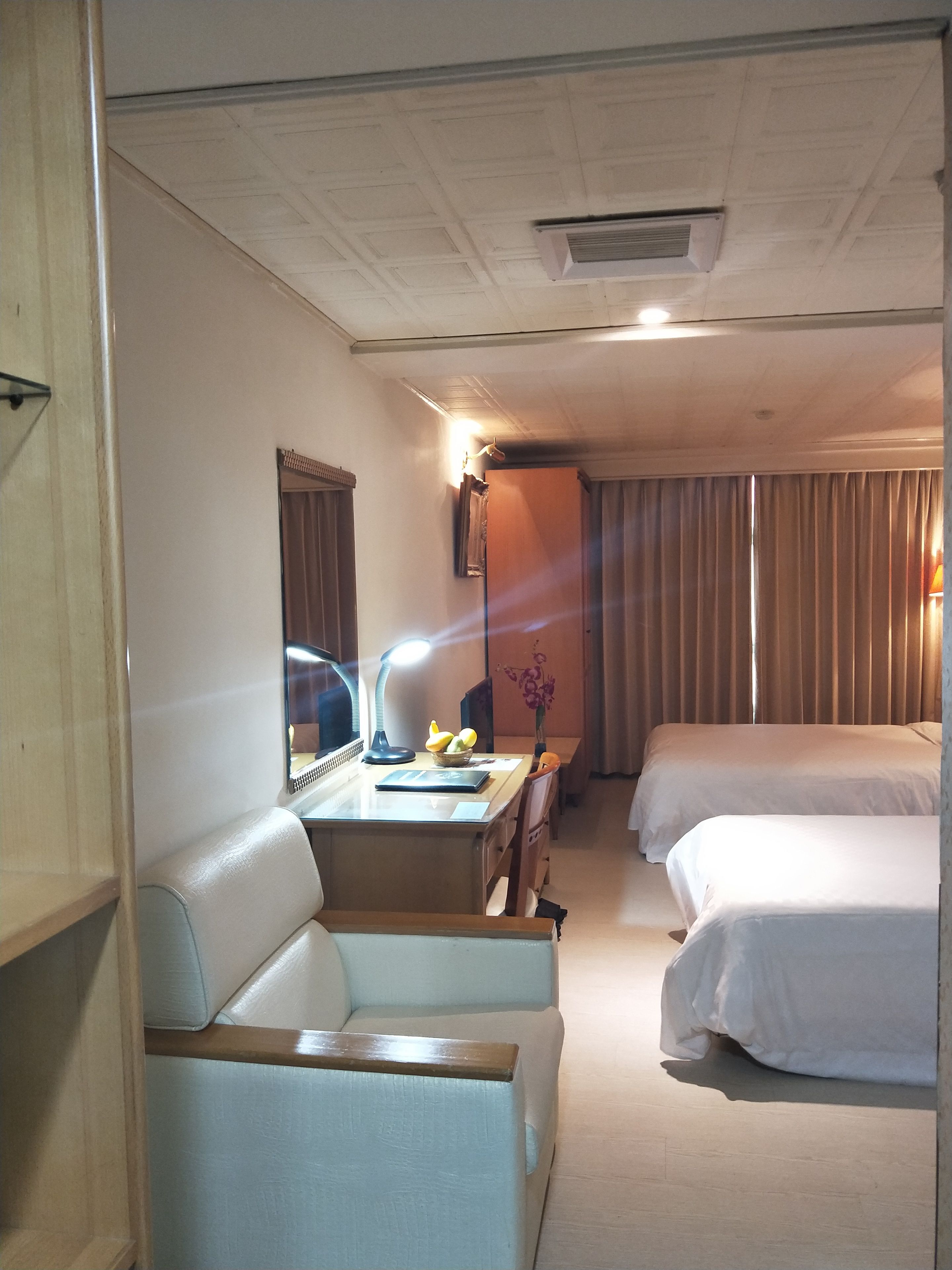 A hotel room with a desk, two beds, a chair, a mirror, and a lamp.