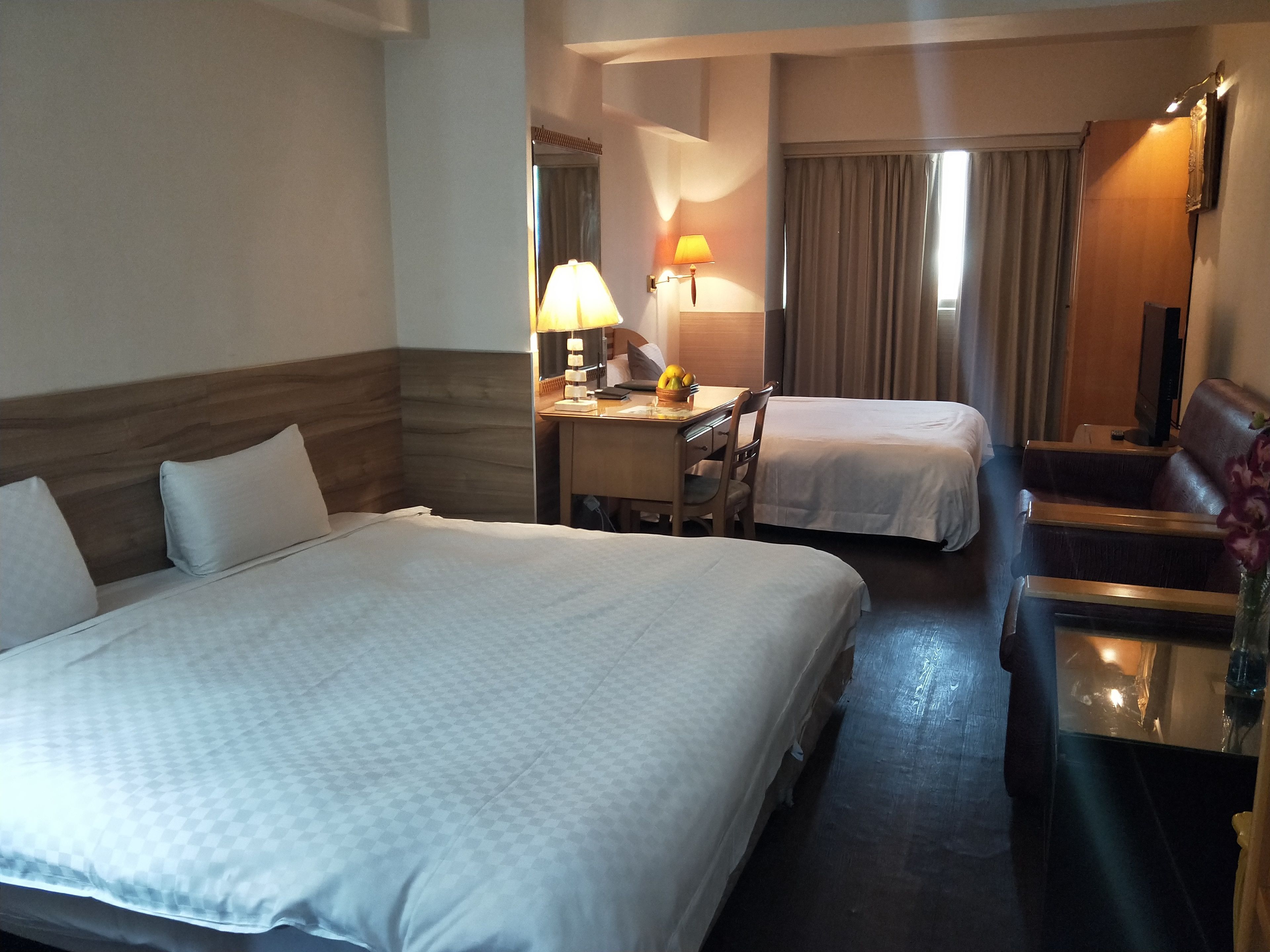 A hotel room with a large bed, a smaller bed, a desk with a lamp, a chair, a sofa, and a mirror.
