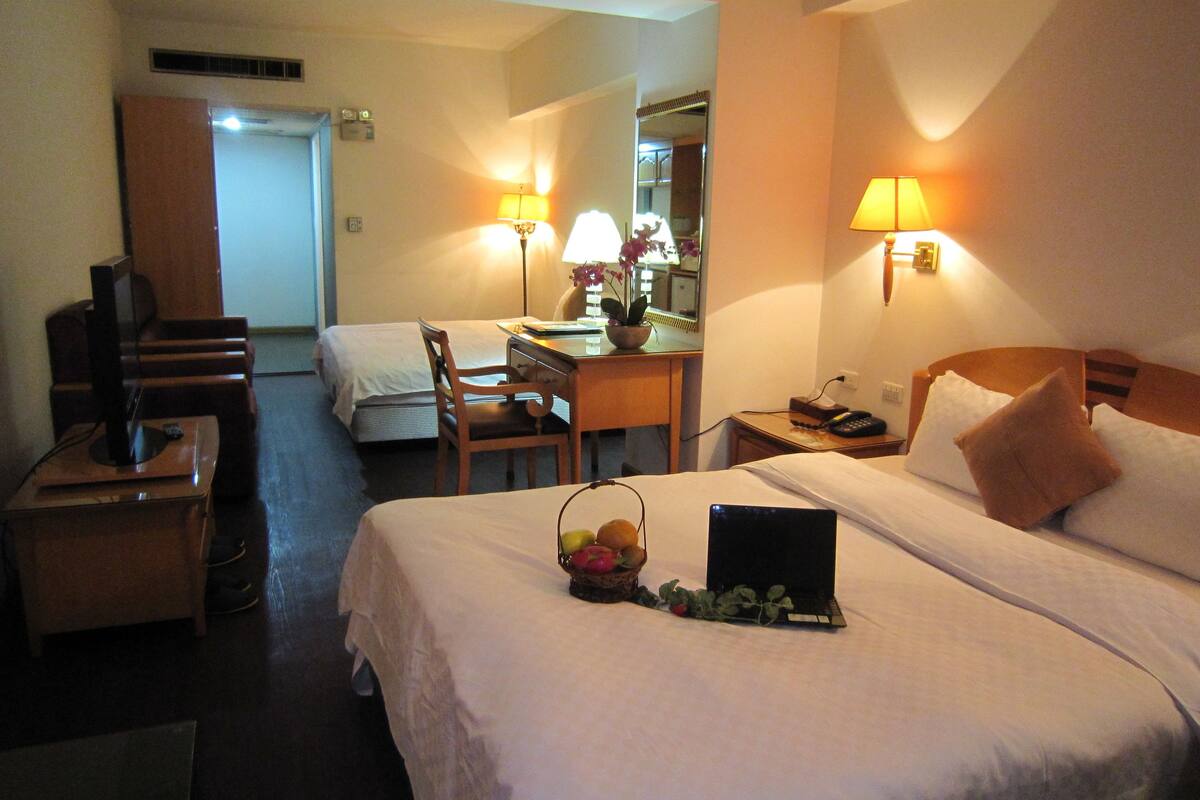 A hotel room with two beds, a desk, a chair, a TV, and a lamp.